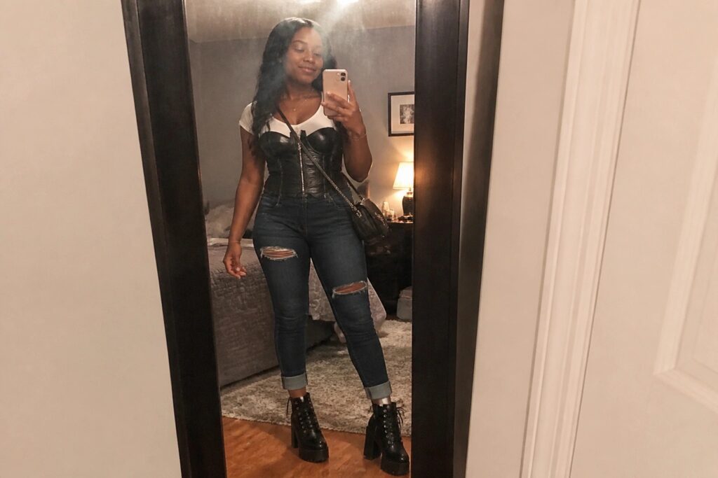 18 Stylish Concert Corset Outfit Ideas for Black Women That Stand Out Woman in white short sleeve tee under black leather corset top, light blue ripped skinny jeans, black combat boots, and small black crossbody bag taking mirror selfie in bedroom.
