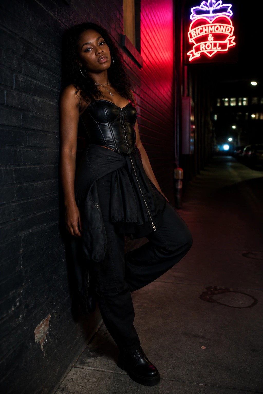 Curly-haired woman in black corset and cargo pants leaning on brick wall under red neon sign