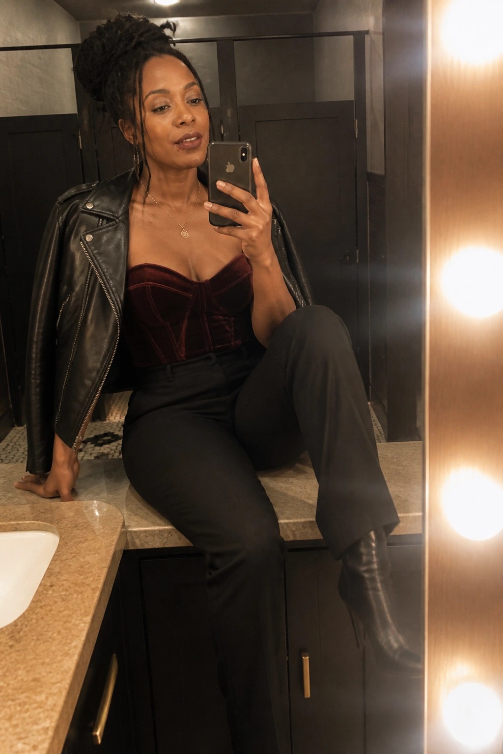 Woman in maroon velvet corset and leather jacket taking mirror selfie in bathroom