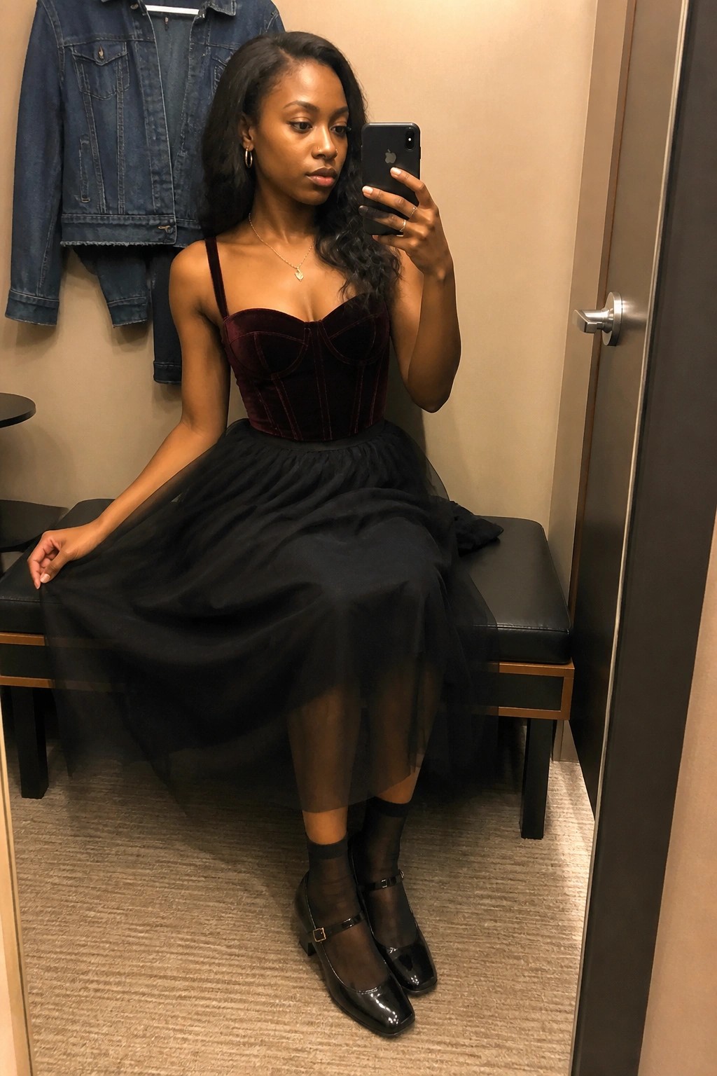 Woman in maroon velvet corset and black tulle skirt takes mirror selfie on bench.