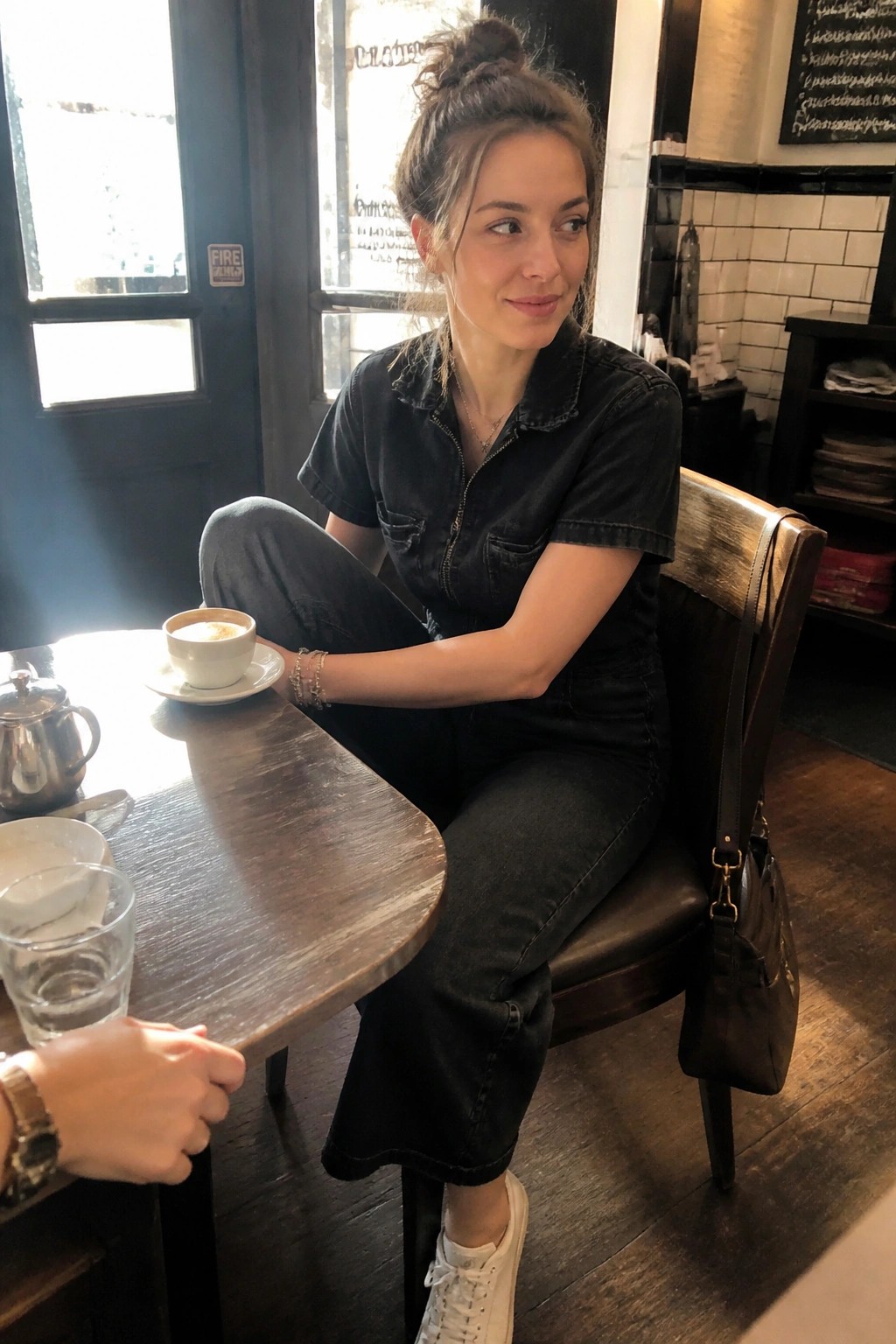 Woman in black denim jumpsuit at wooden cafe table with coffee cup