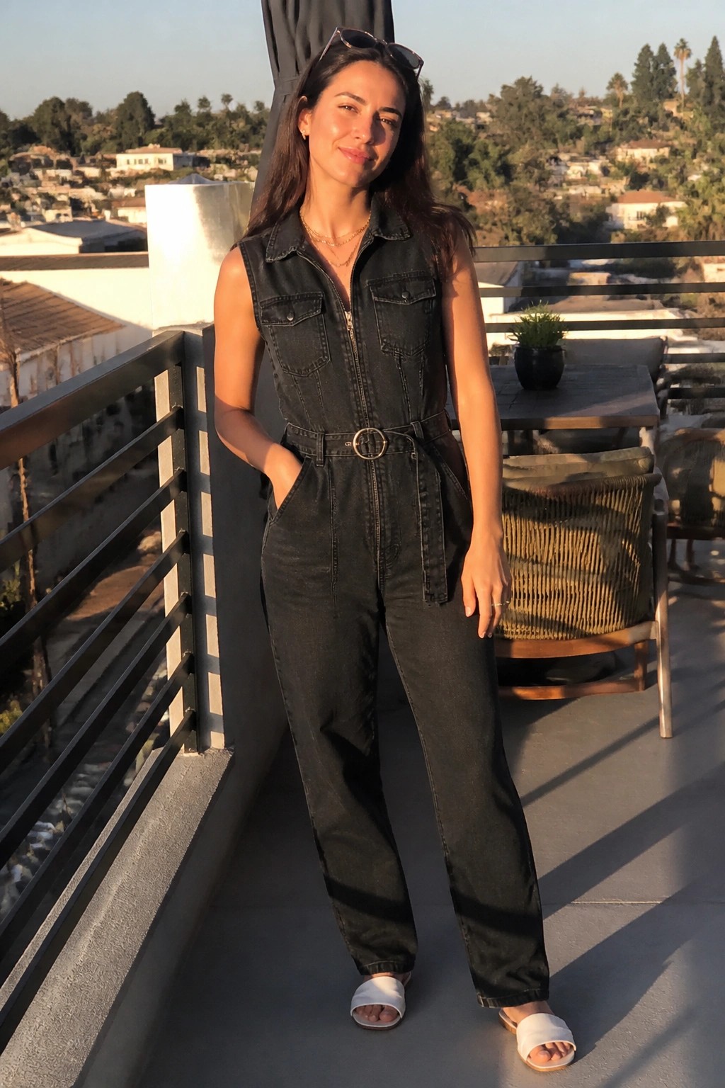 Woman in black sleeveless denim jumpsuit on sunny balcony overlooking hills and homes.