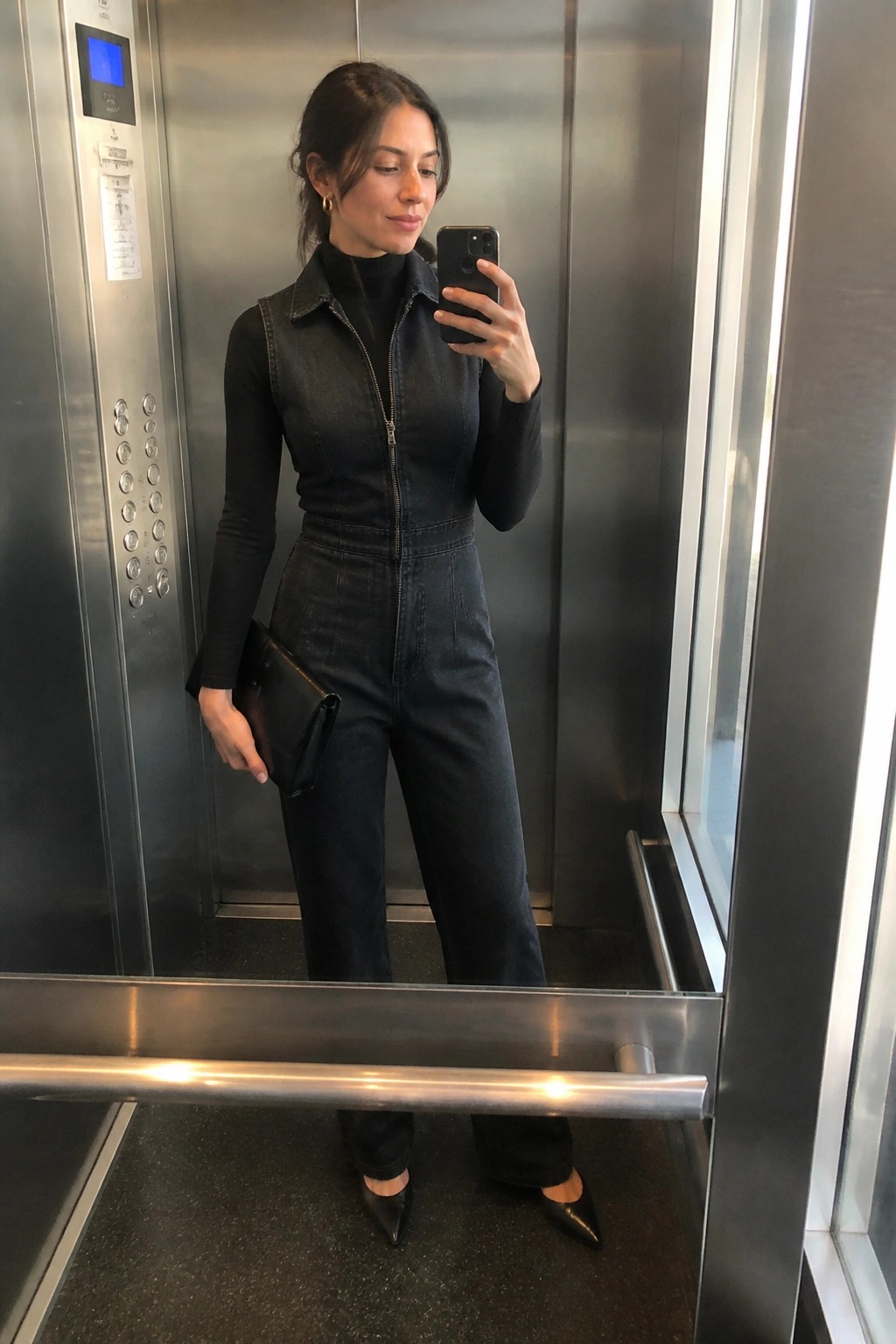 Woman in elevator mirror selfie wearing black denim jumpsuit zipped over black turtleneck top with black pointed flats and black clutch in hand.