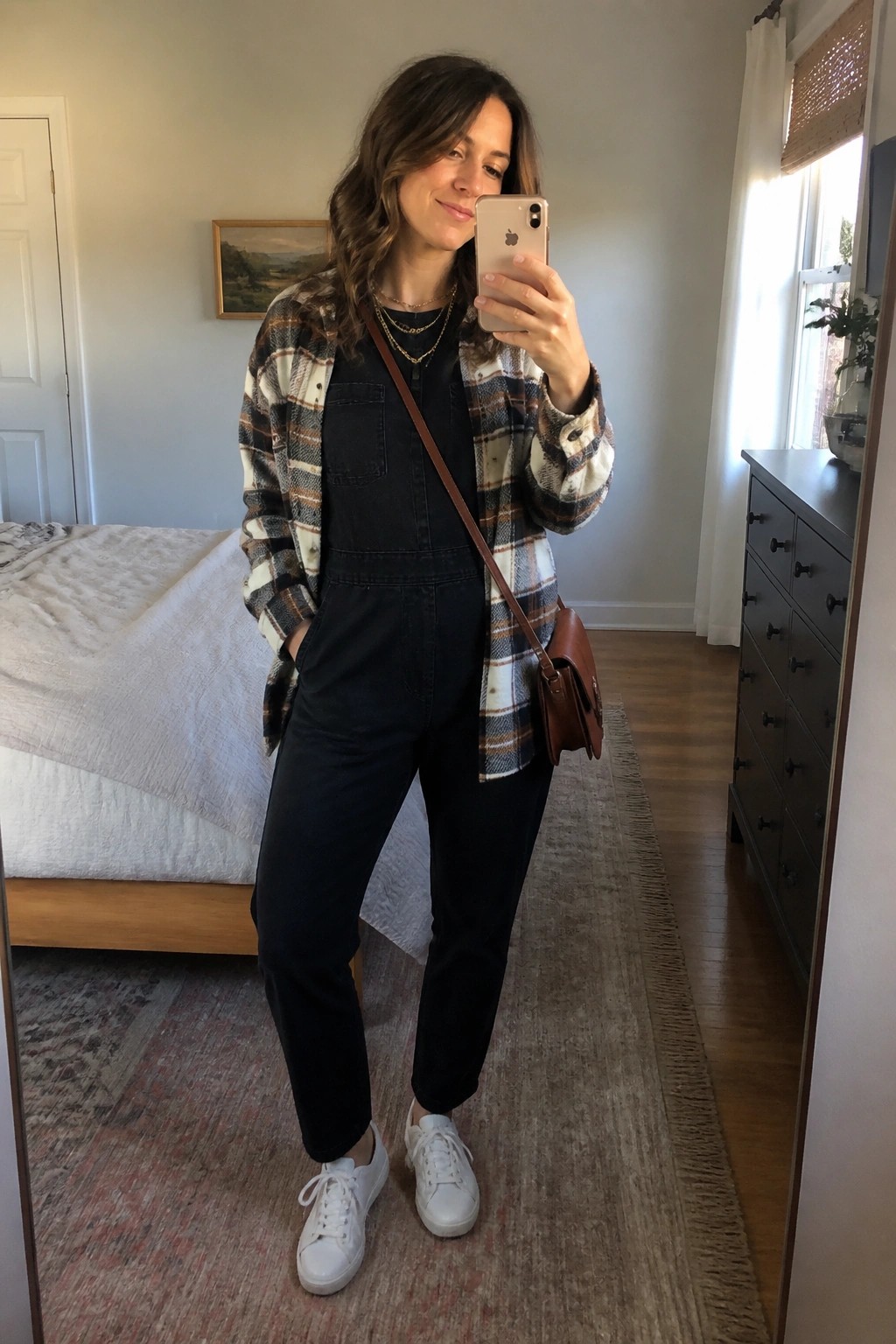 Woman in plaid shirt over black jumpsuit taking mirror selfie in bedroom
