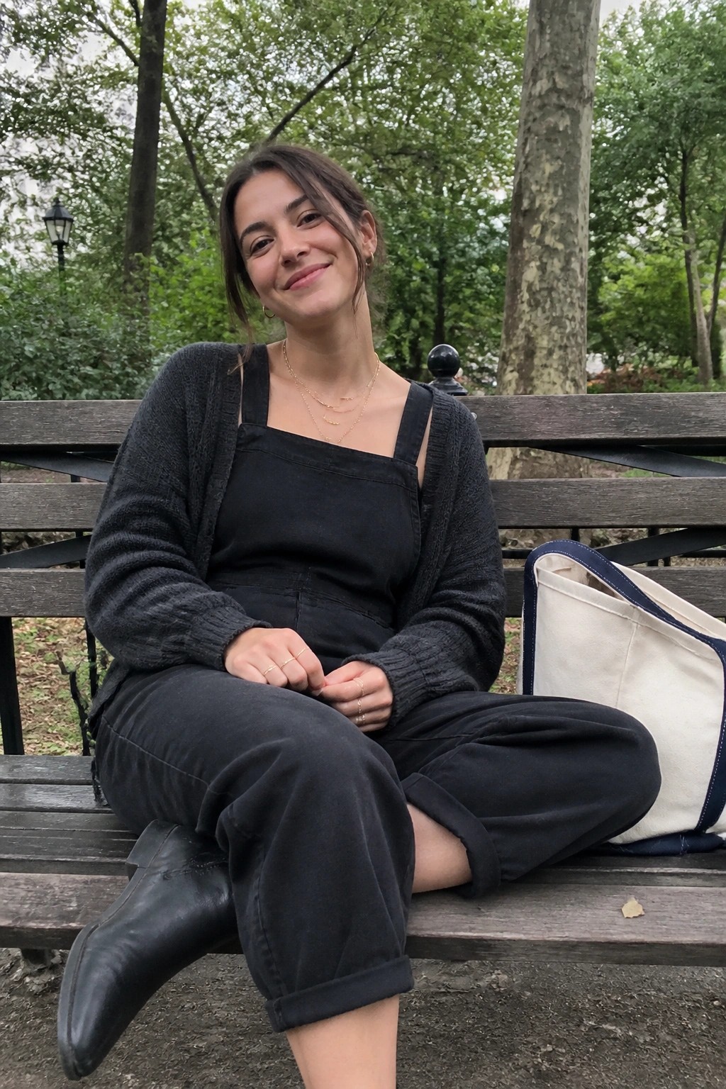Smiling woman in black cardigan and overalls on park bench with tote bag.