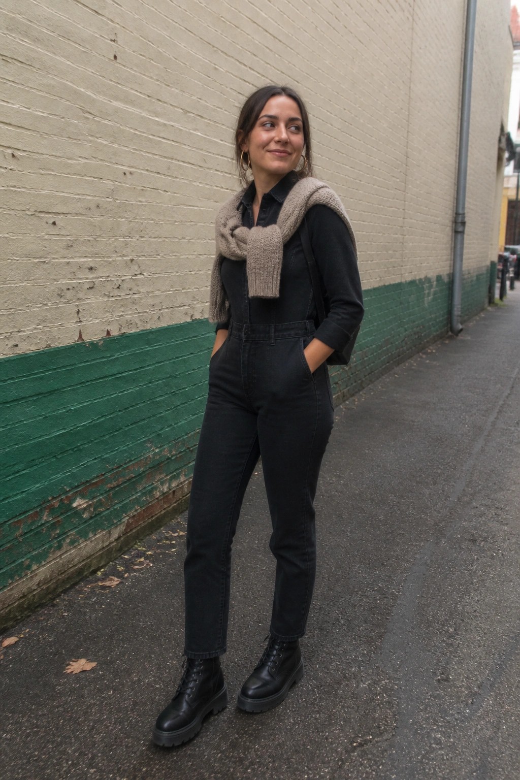 Woman in black overalls with beige scarf and boots stands in alley.