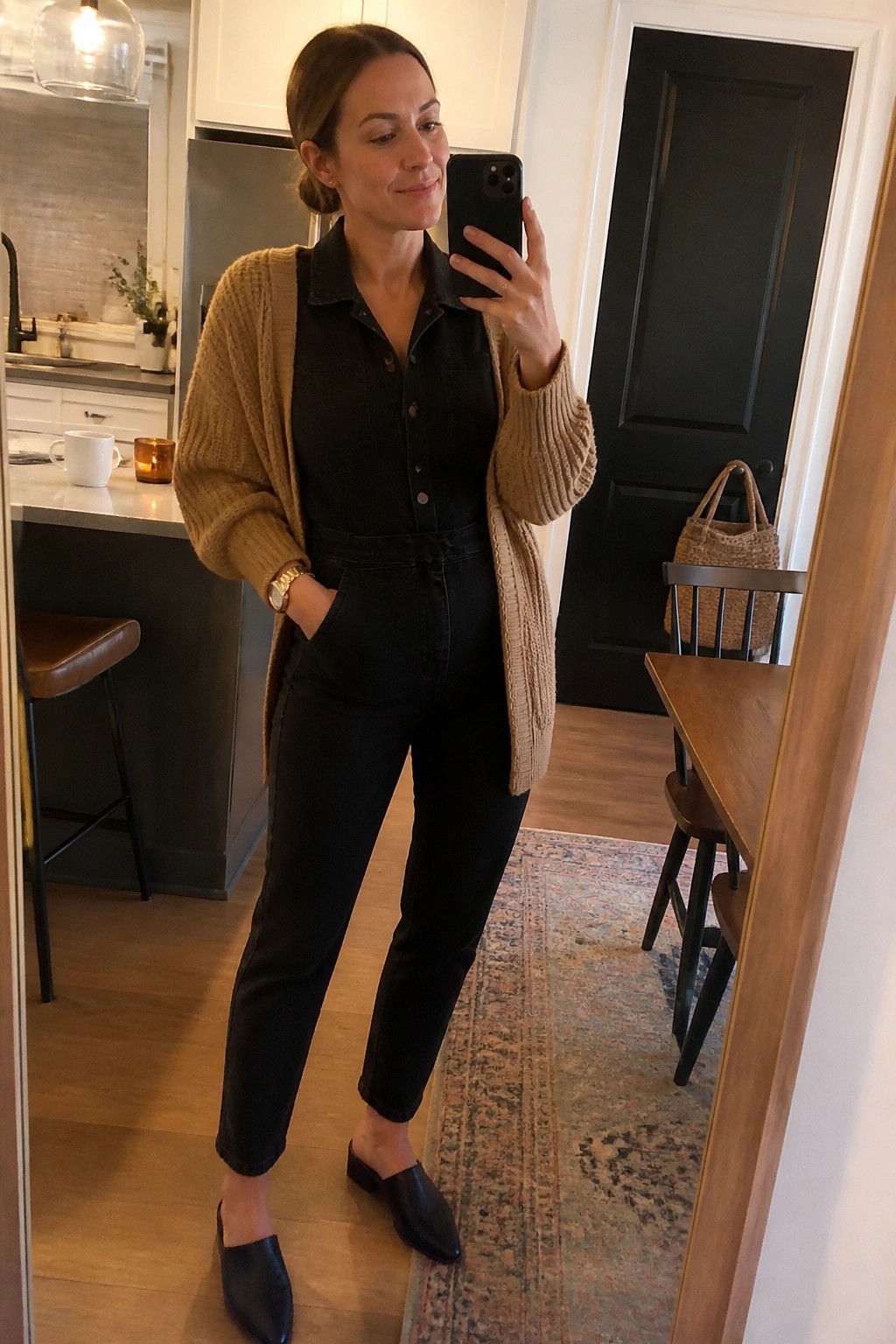 Woman in black denim jumpsuit and beige cardigan taking mirror selfie in kitchen