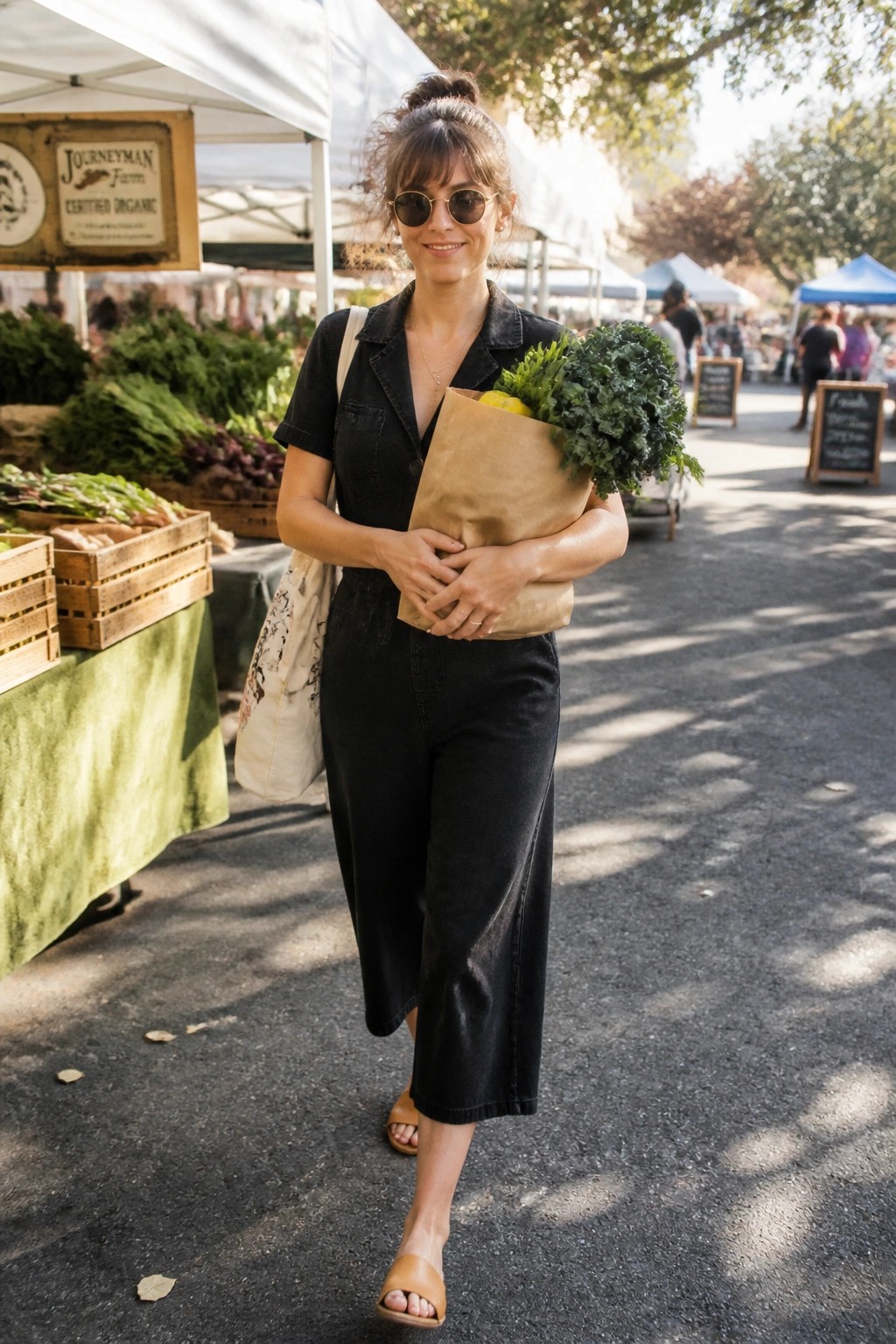 Woman in black short-sleeve denim jumpsuit holding a paper grocery bag with greens and lemons, wearing round sunglasses, canvas tote, and tan slide sandals at an outdoor market.