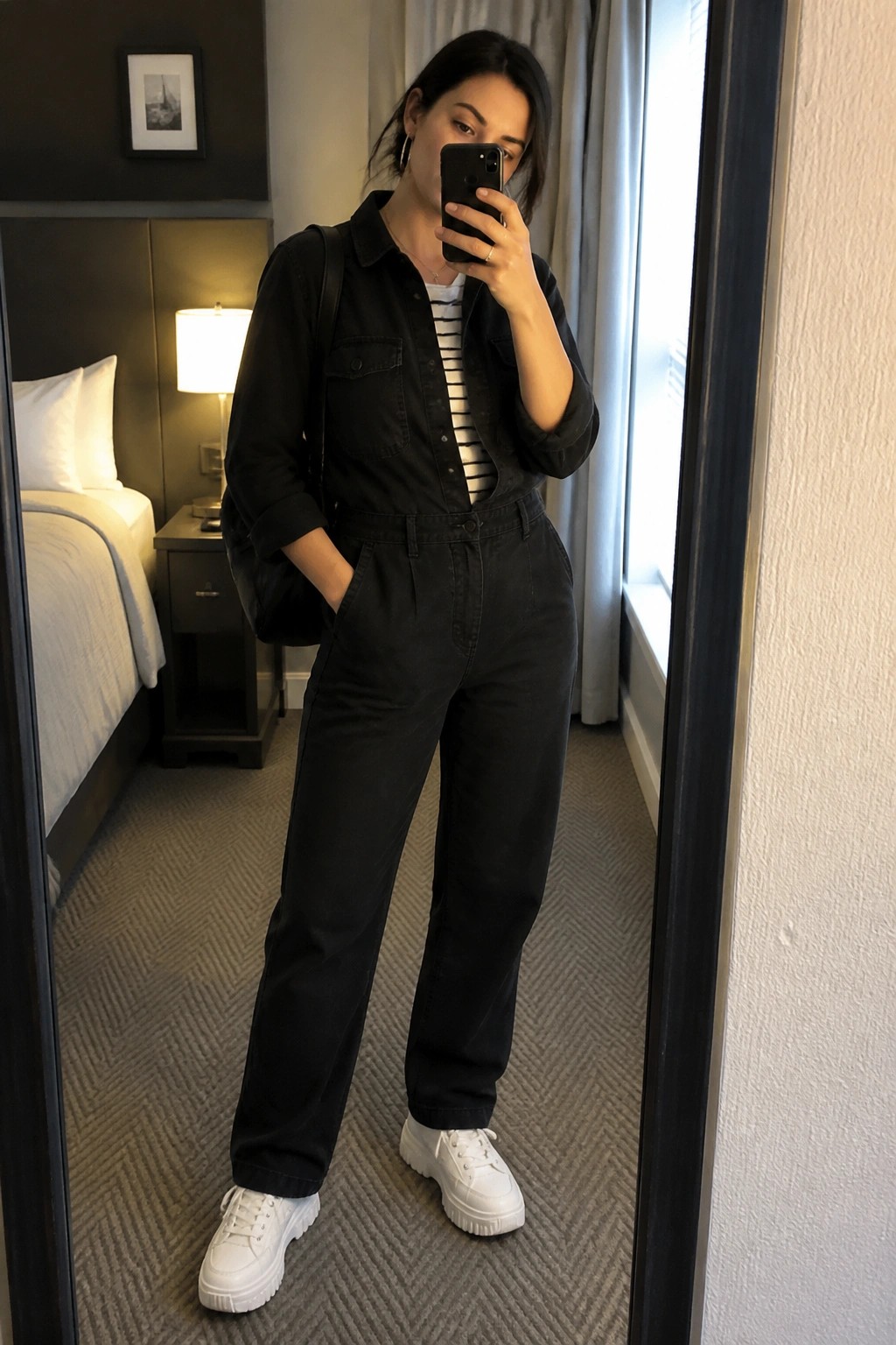 Woman in black jumpsuit over striped top with white sneakers taking mirror selfie in hotel room
