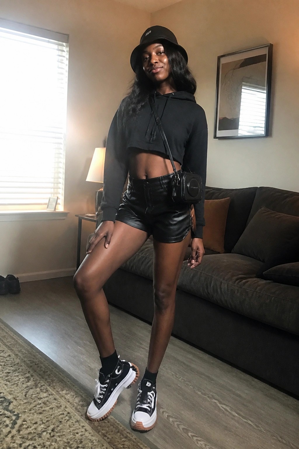 Woman in black cropped hoodie, leather shorts, bucket hat, and platform sneakers in living room