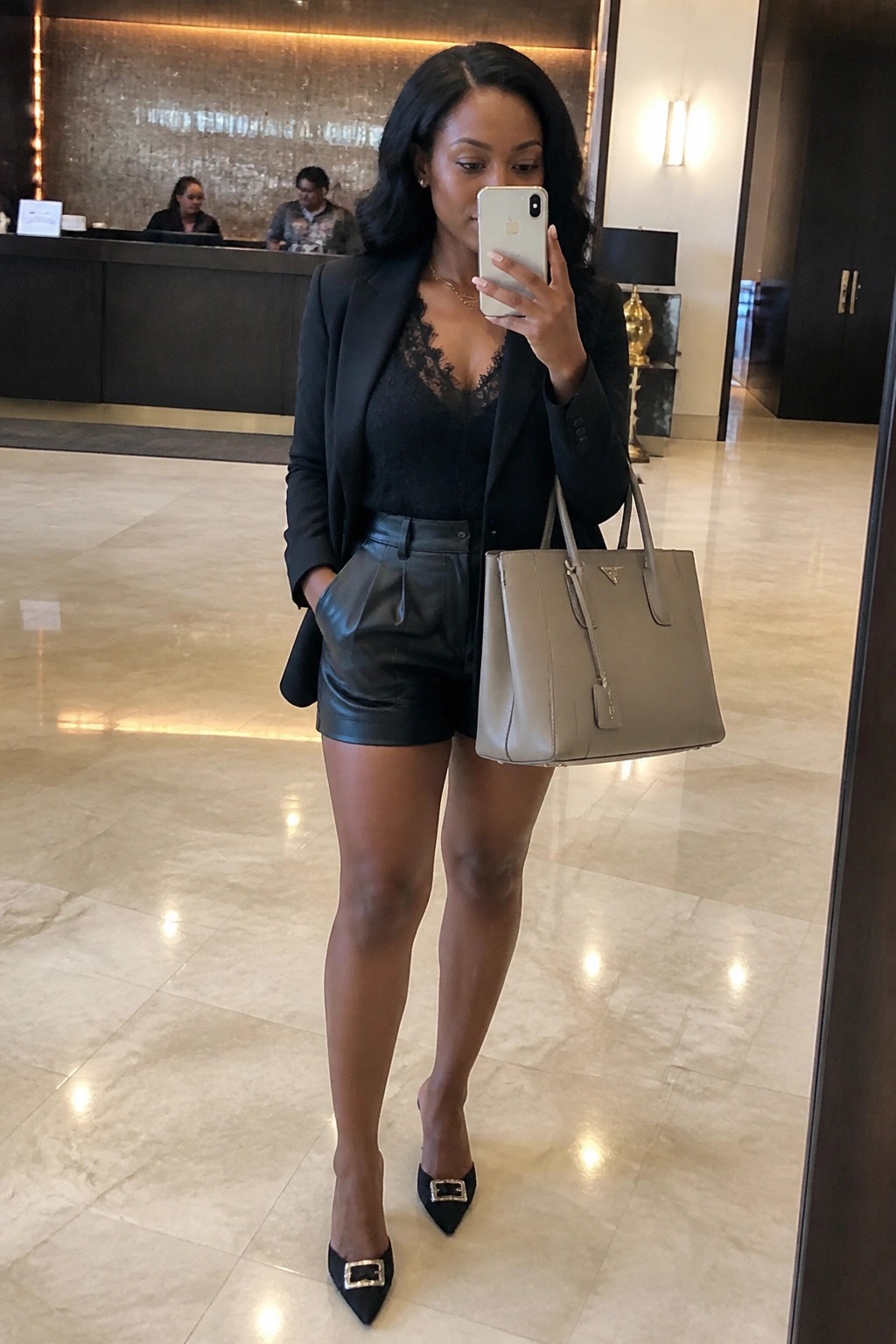 Woman in black blazer, lace top and leather shorts taking mirror selfie with gray handbag.