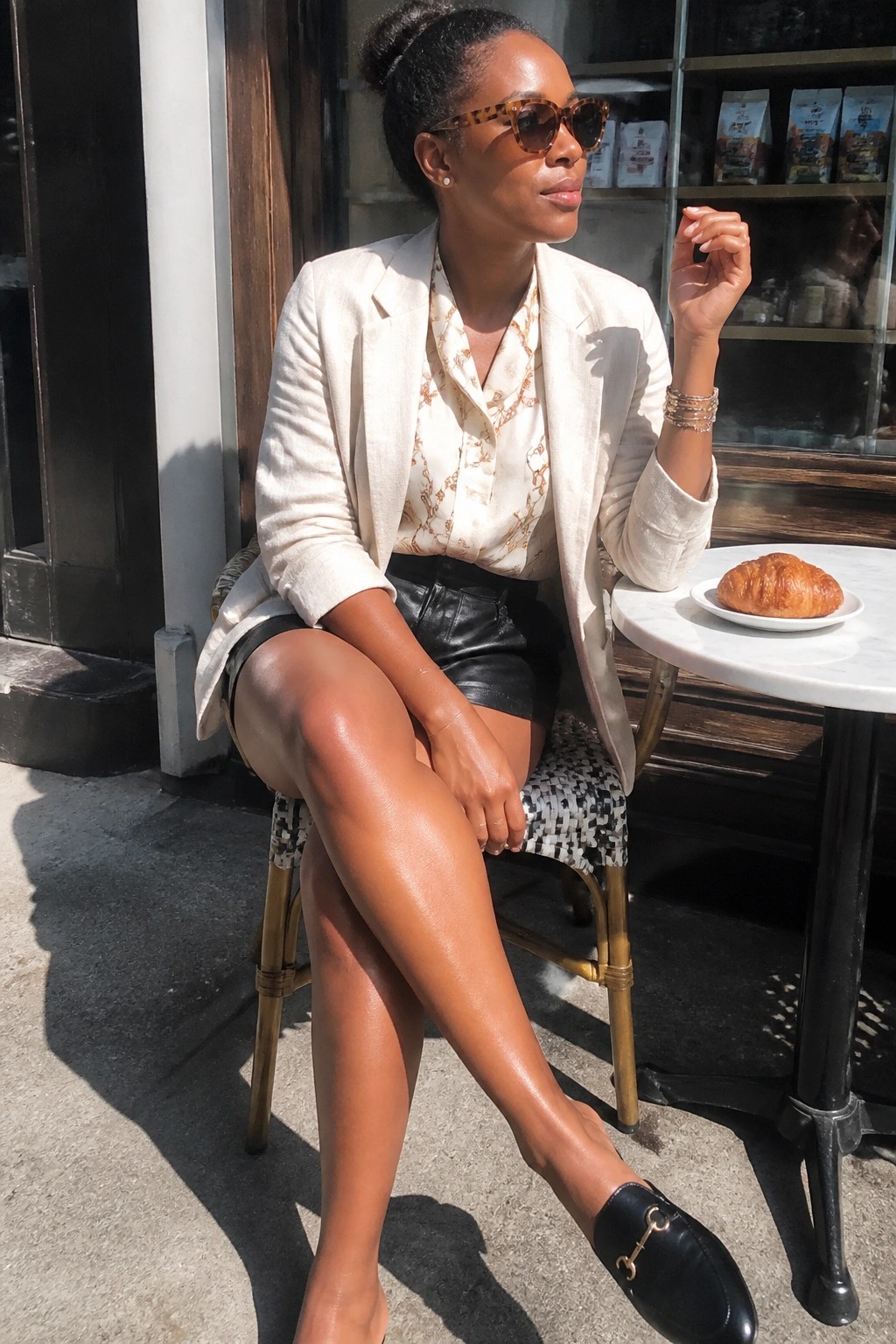 Woman in cream blazer and black leather shorts at sunny cafe table with croissant