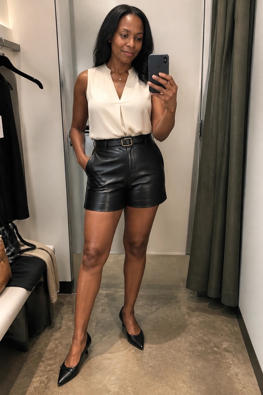 Woman in cream blouse and black leather shorts taking mirror selfie in fitting room