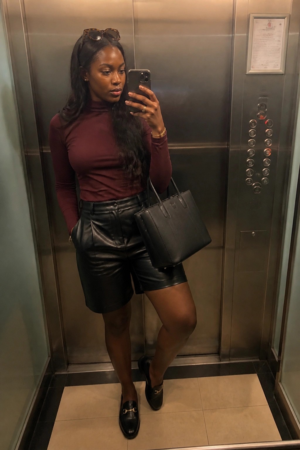 A woman standing in an elevator taking a mirror selfie while wearing black faux leather shorts, a maroon turtleneck top tucked in, black loafers with gold hardware, and carrying a black structured tote bag.