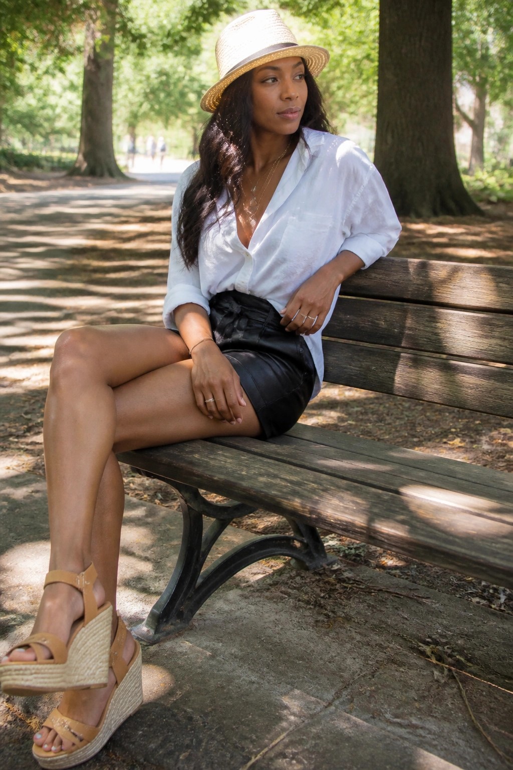 Black woman seated on park bench wearing white button down shirt, black faux leather mini skirt with front tie, layered gold necklaces, straw hat, and tan wedge espadrille sandals.