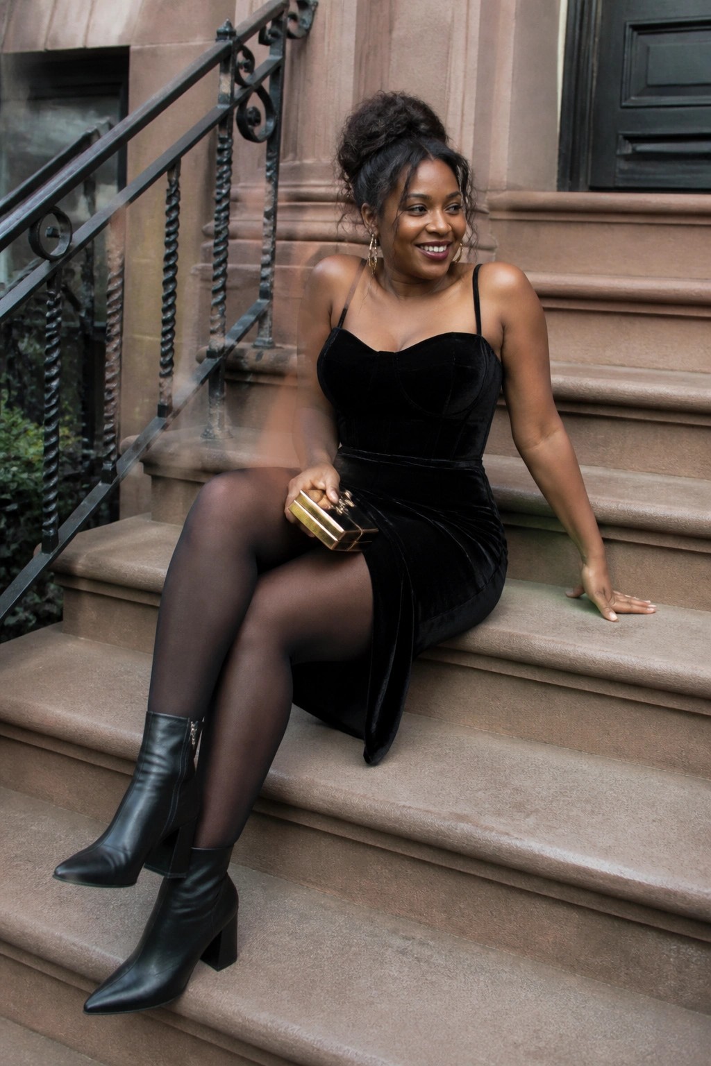 Woman in black velvet dress and boots sits on stone steps holding gold clutch