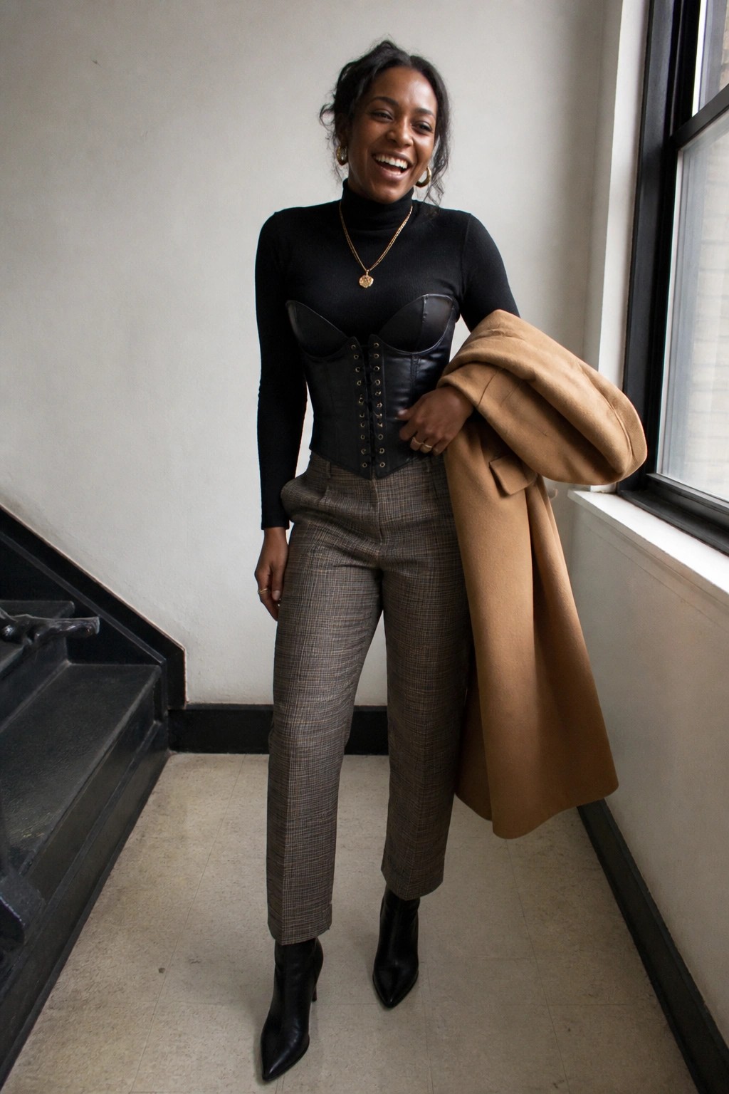 Woman wearing a black turtleneck under a black leather corset with brown plaid trousers and black ankle boots while holding a camel coat over one arm.