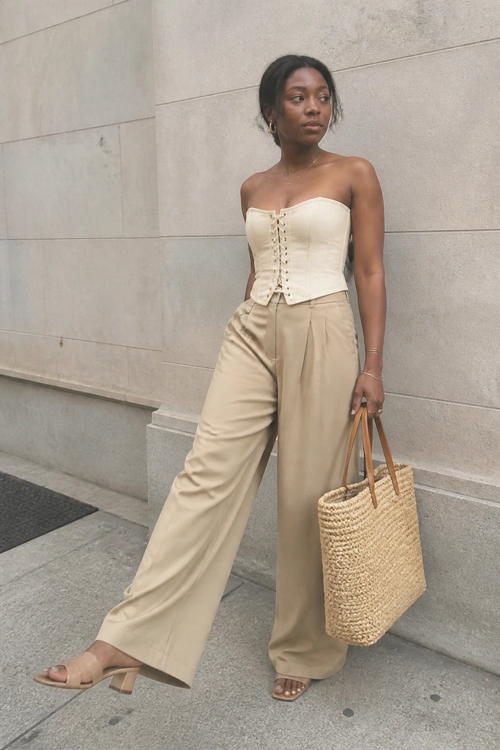 Woman in beige corset top and wide-leg trousers holding woven tote by stone wall.