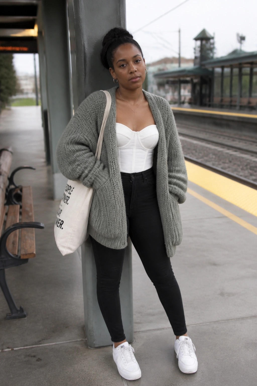 Black woman in gray knit cardigan over white corset at train platform with tote bag.