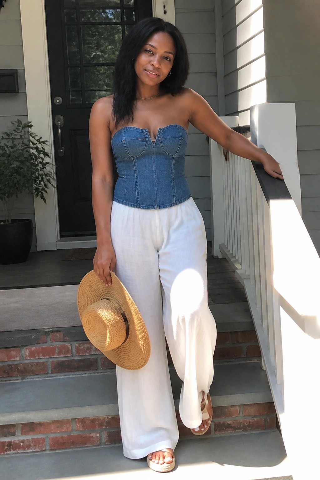 Woman in blue denim corset top and white wide-leg pants holding straw hat on steps