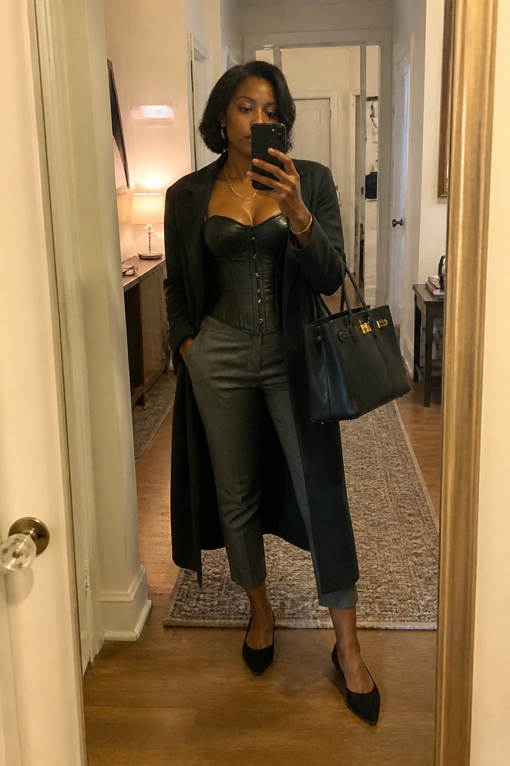 Woman in black leather corset, gray pants, and coat taking mirror selfie with handbag