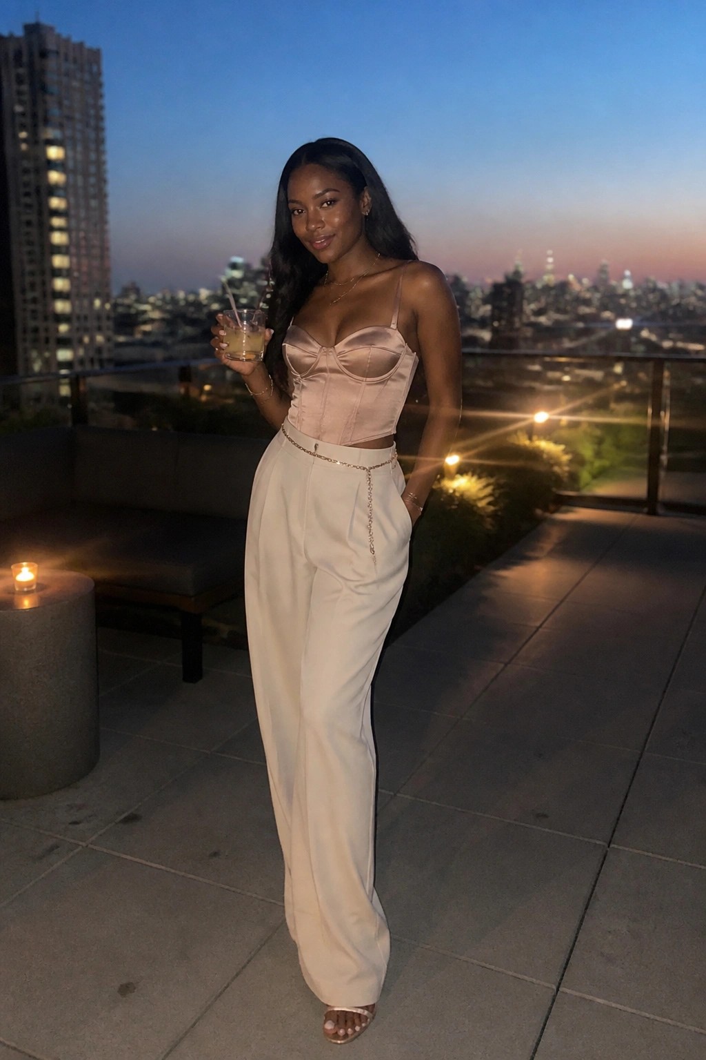 Woman in pink satin corset and white pants holding drink on city rooftop at dusk