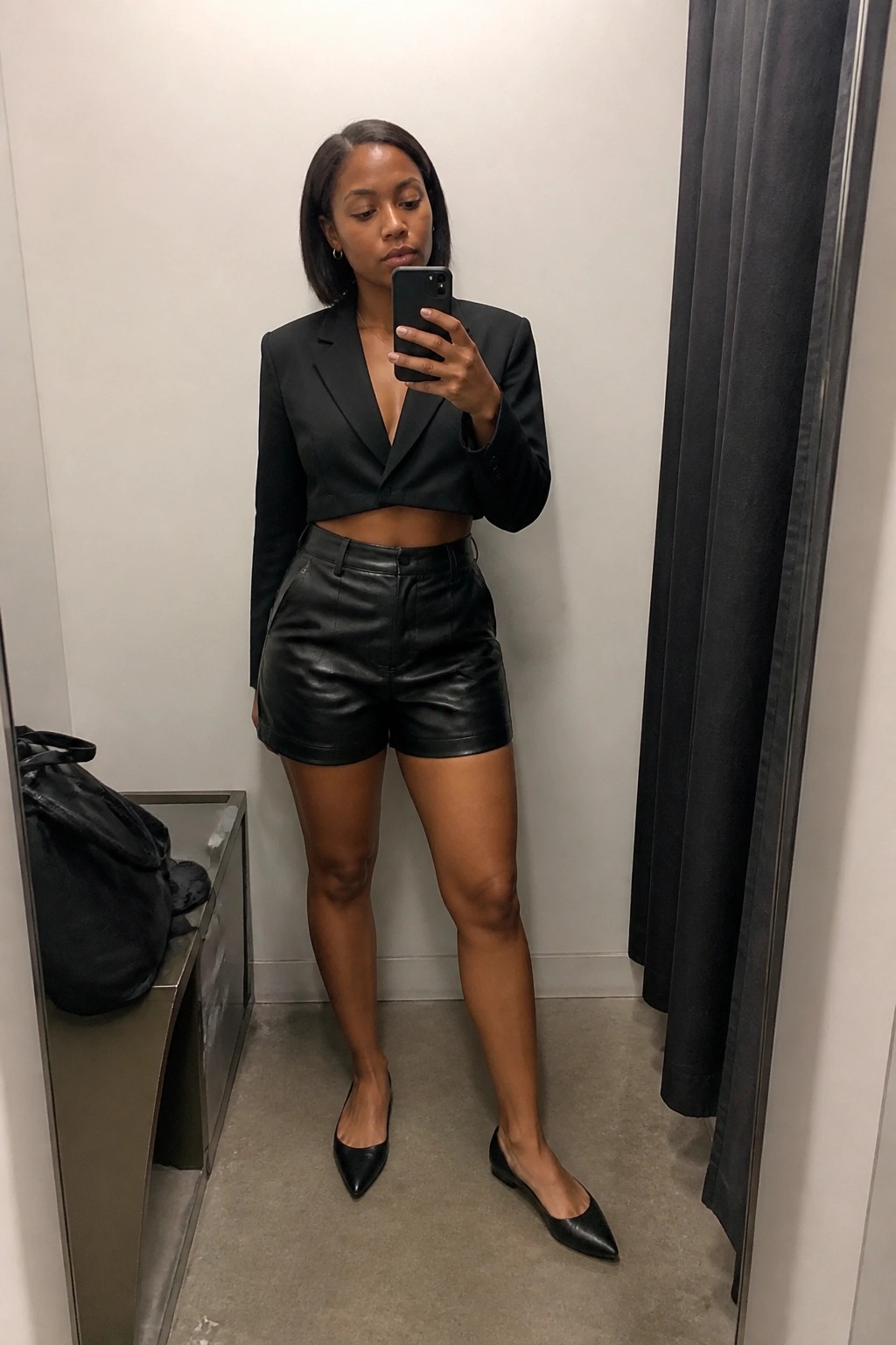 Woman in black cropped blazer and leather shorts taking mirror selfie in fitting room