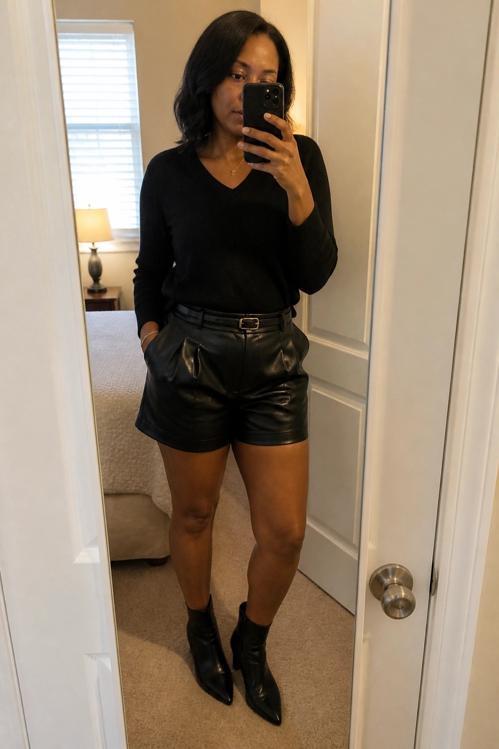Woman in black sweater and leather shorts takes mirror selfie wearing ankle boots