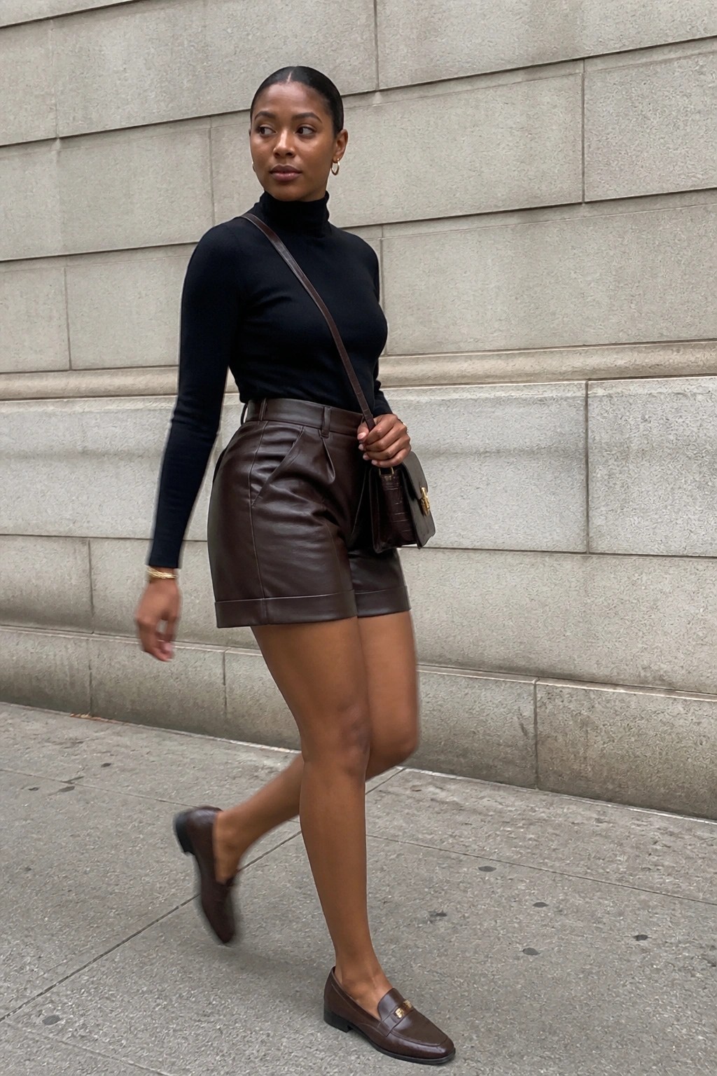 Woman in black turtleneck and brown leather shorts walking on city sidewalk with handbag