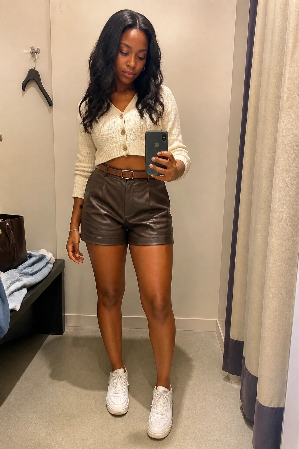 Woman in white cropped cardigan and brown leather shorts taking mirror selfie in fitting room
