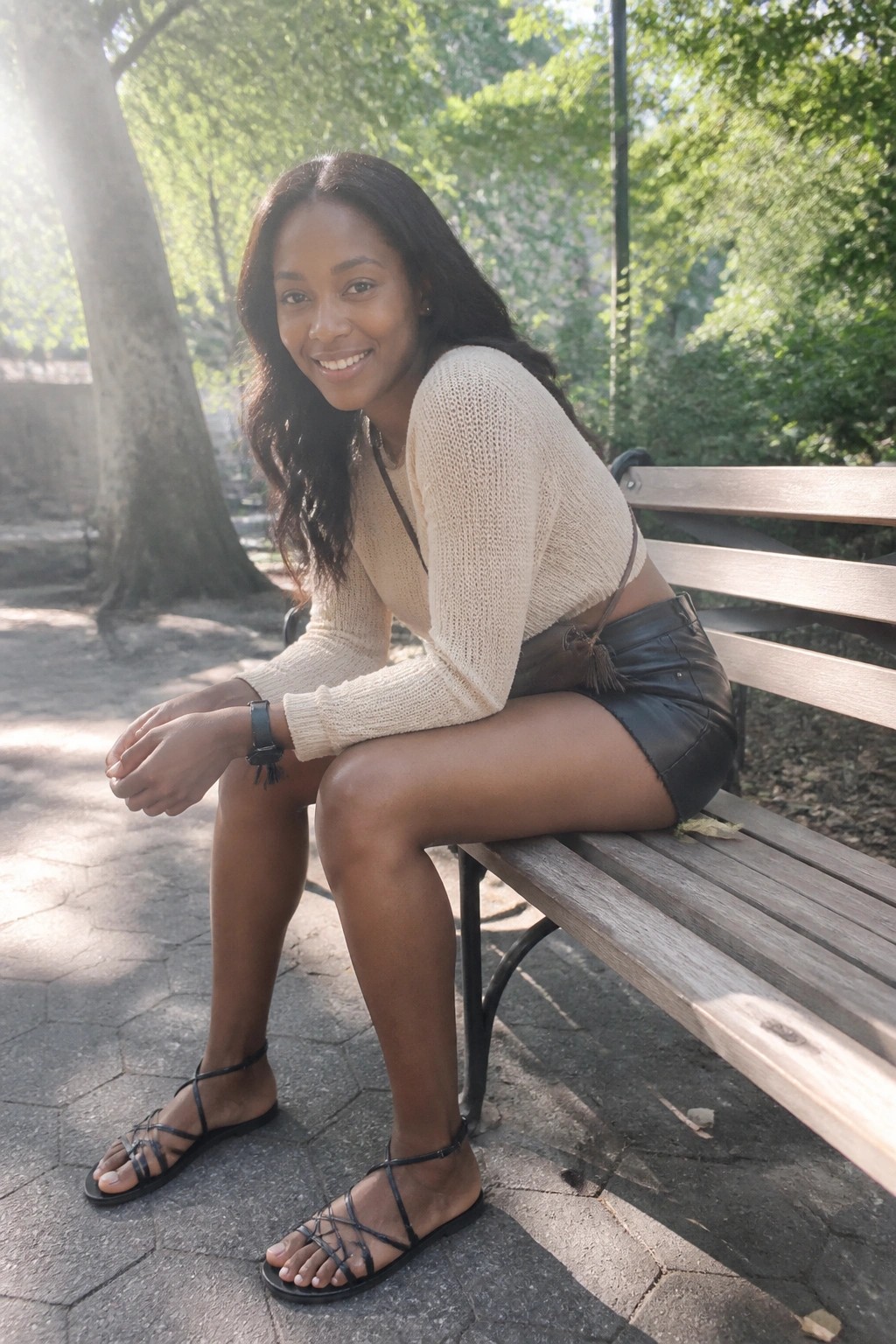 Smiling woman in cream knit sweater and black leather shorts on sunny park bench.