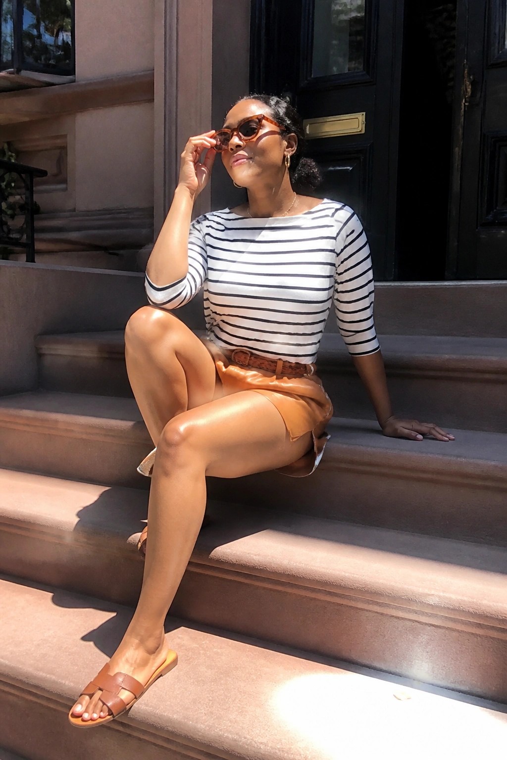 Woman in striped top and leather shorts sitting on stone steps adjusting sunglasses