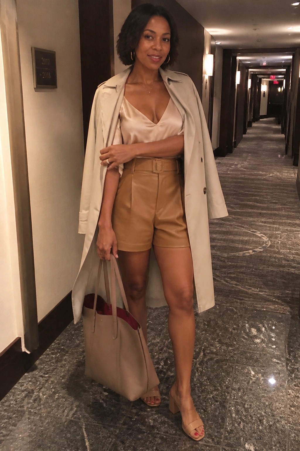 A Black woman in tan leather shorts, beige trench coat over a satin camisole, holding a large tote bag, standing in a hallway.