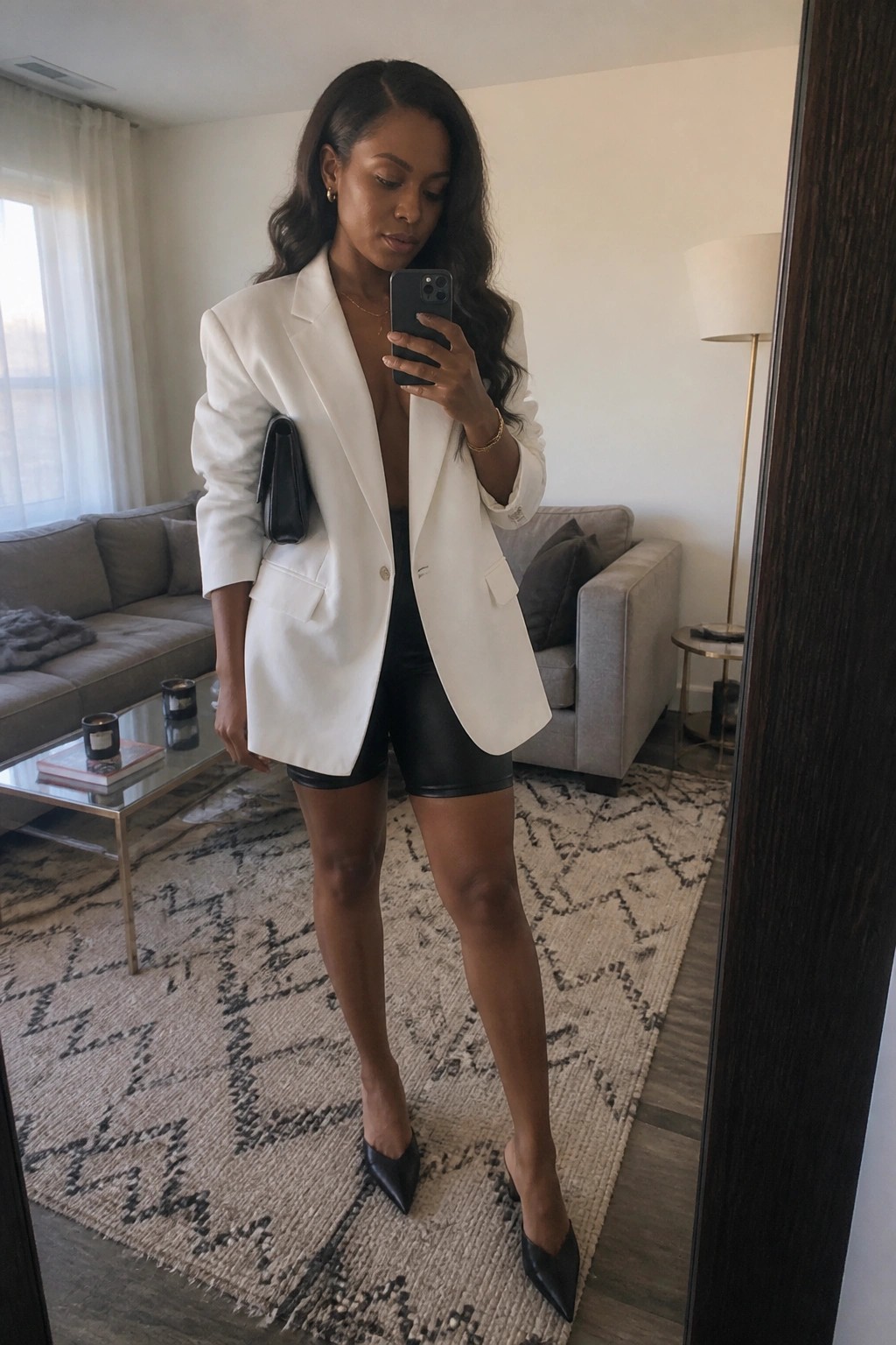 Woman in oversized white blazer and black shorts taking mirror selfie in living room