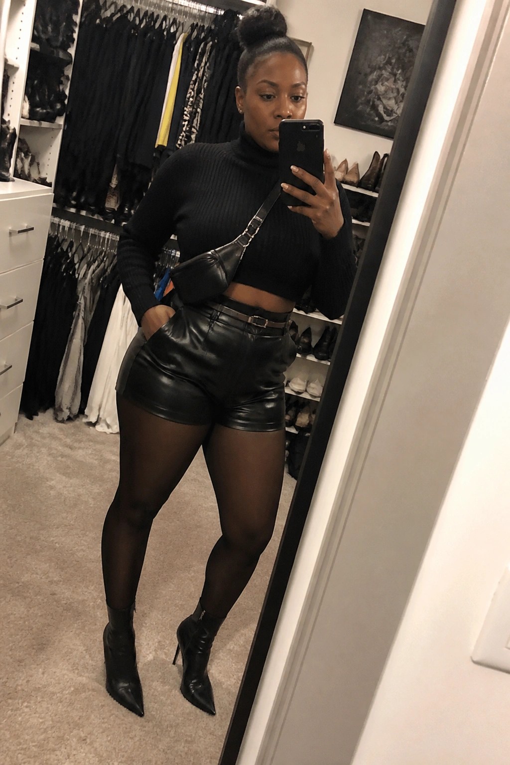Woman in black crop top and leather shorts taking mirror selfie in closet.