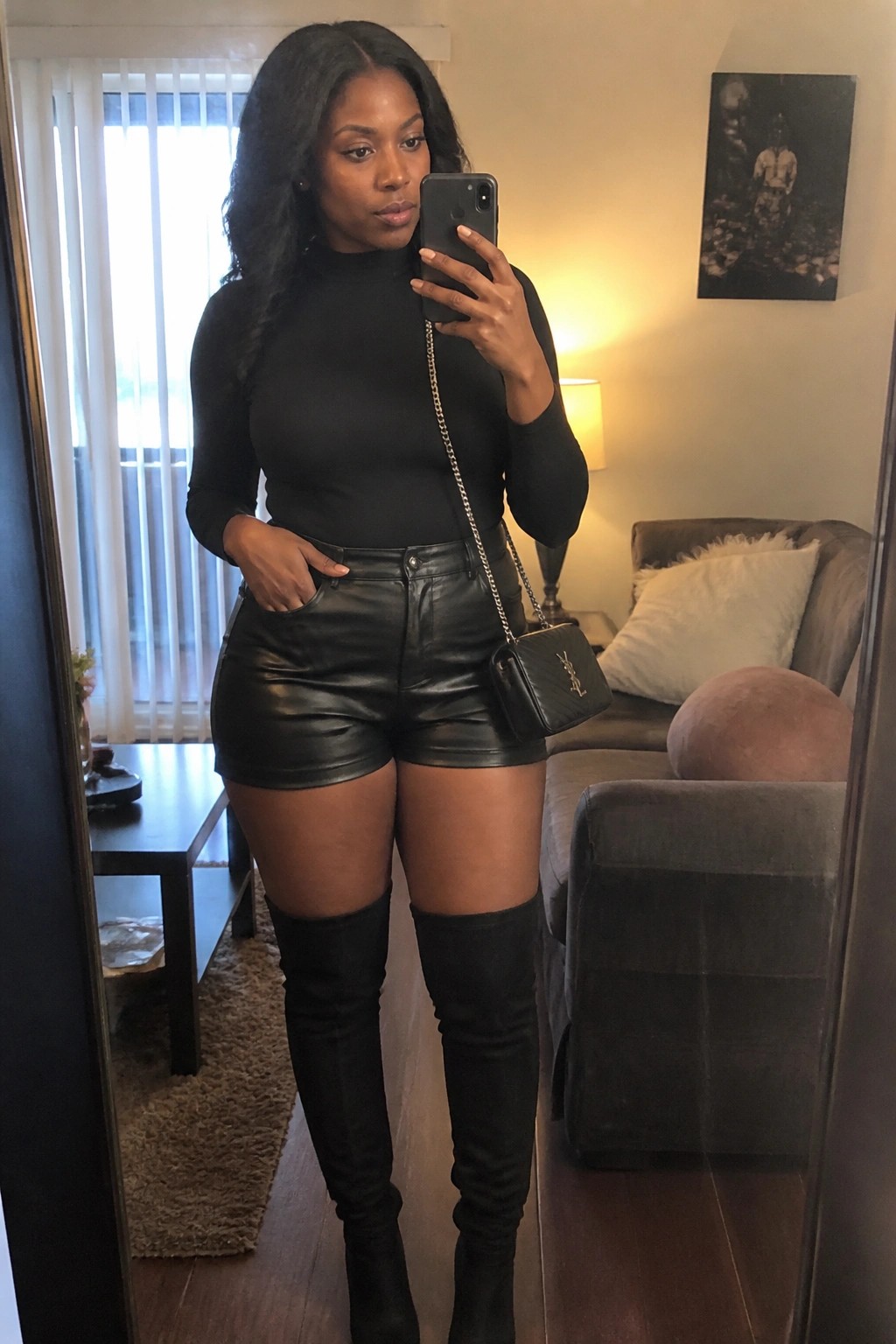 Woman in black turtleneck, leather shorts, and thigh-high boots holding phone with YSL bag