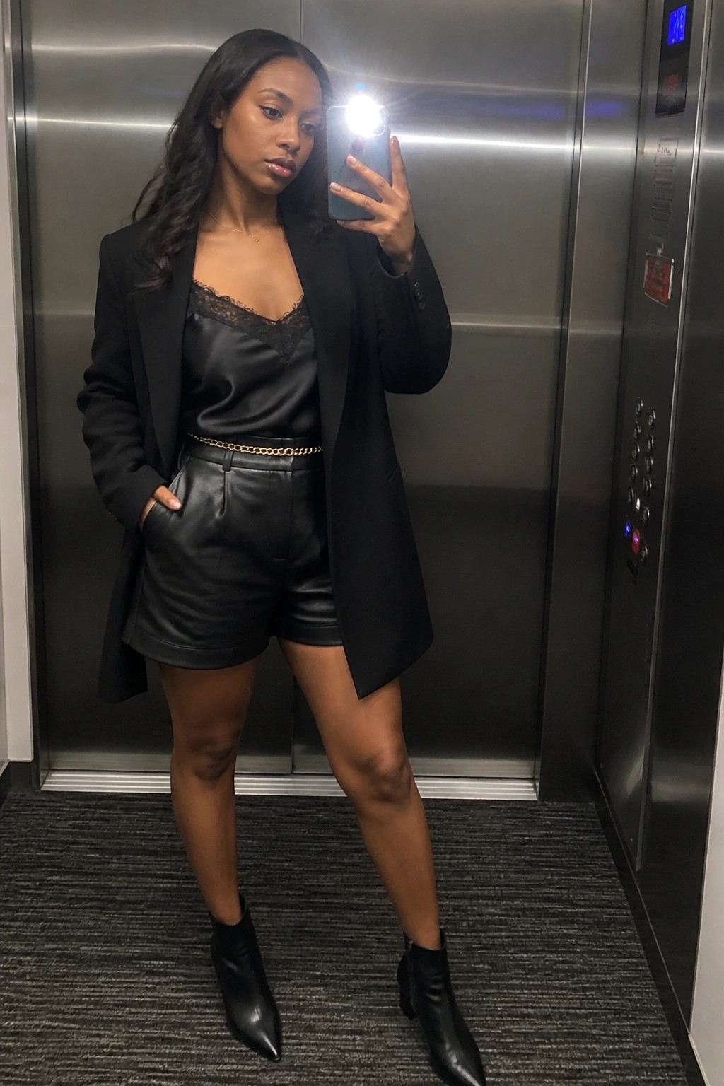 Woman in black blazer and leather shorts taking selfie in elevator