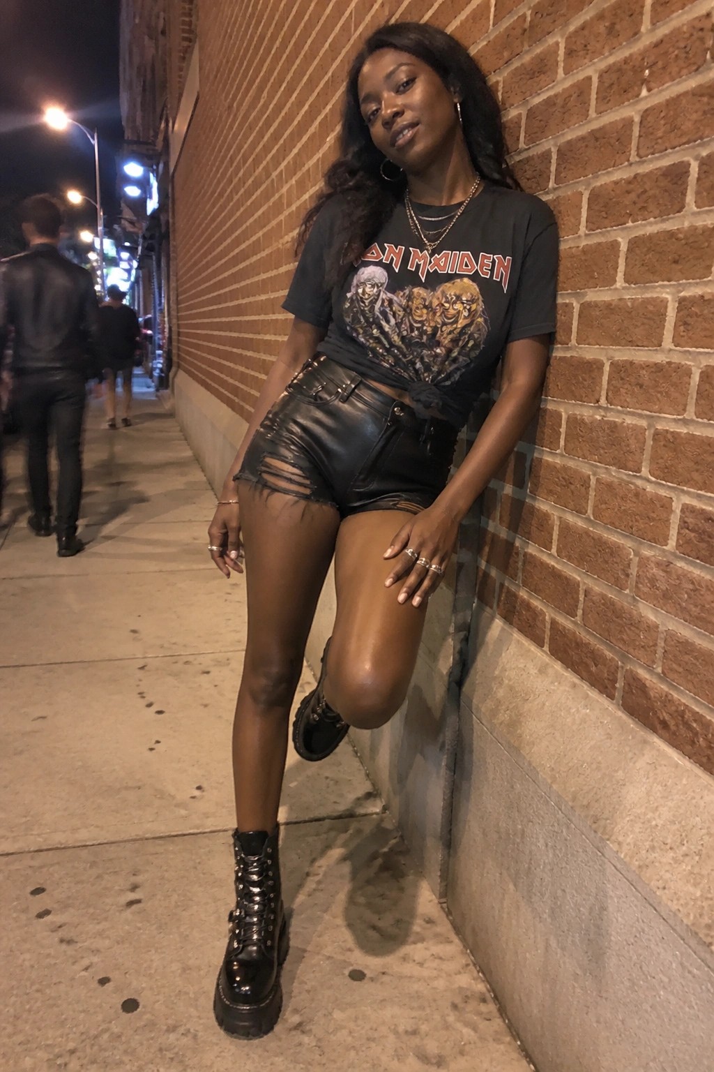 Woman in Iron Maiden t-shirt and ripped leather shorts leans on brick wall at night.