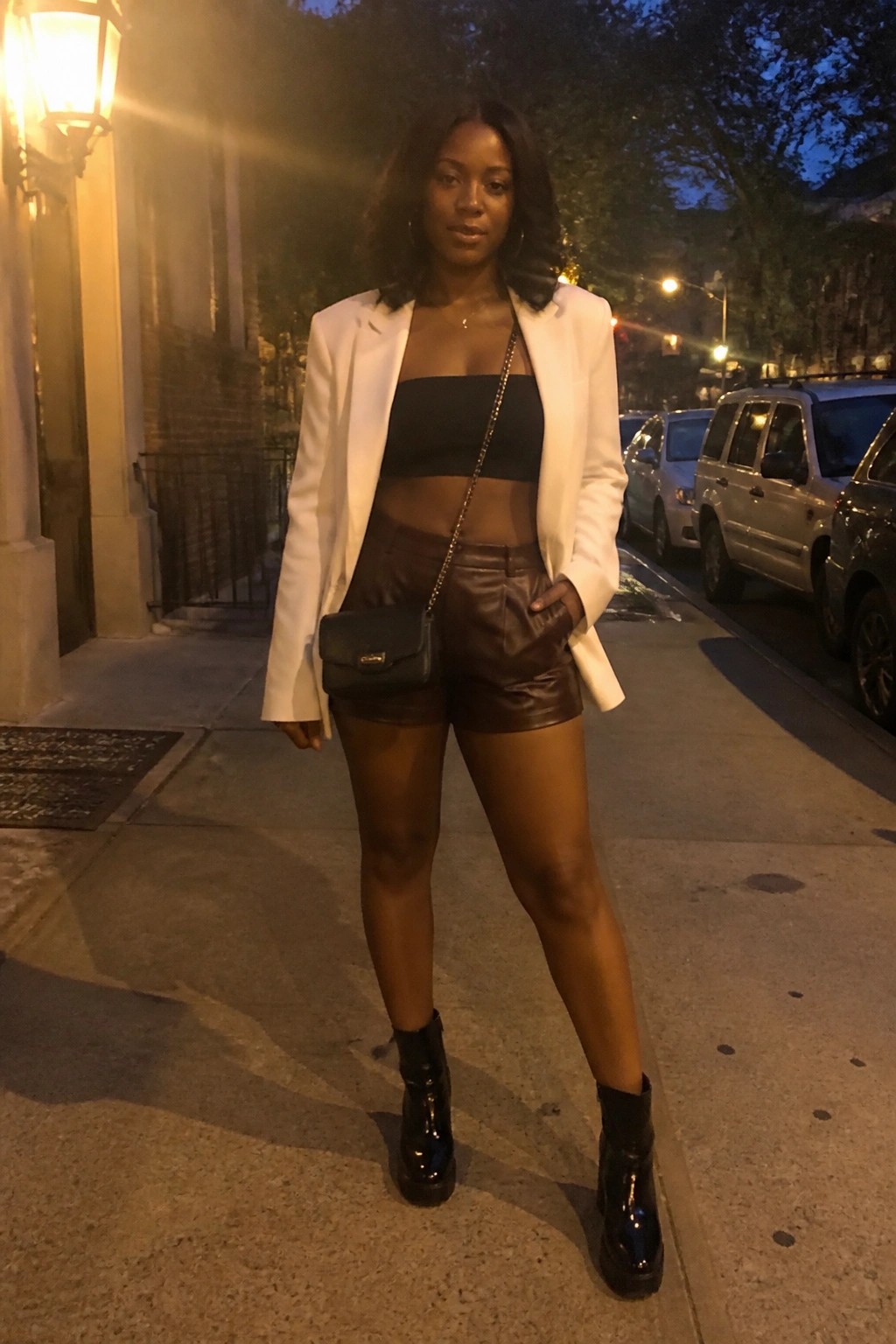 A Black woman wearing brown leather shorts, a black crop top, an open white blazer, black patent boots, and a black crossbody bag standing on a sidewalk at night.