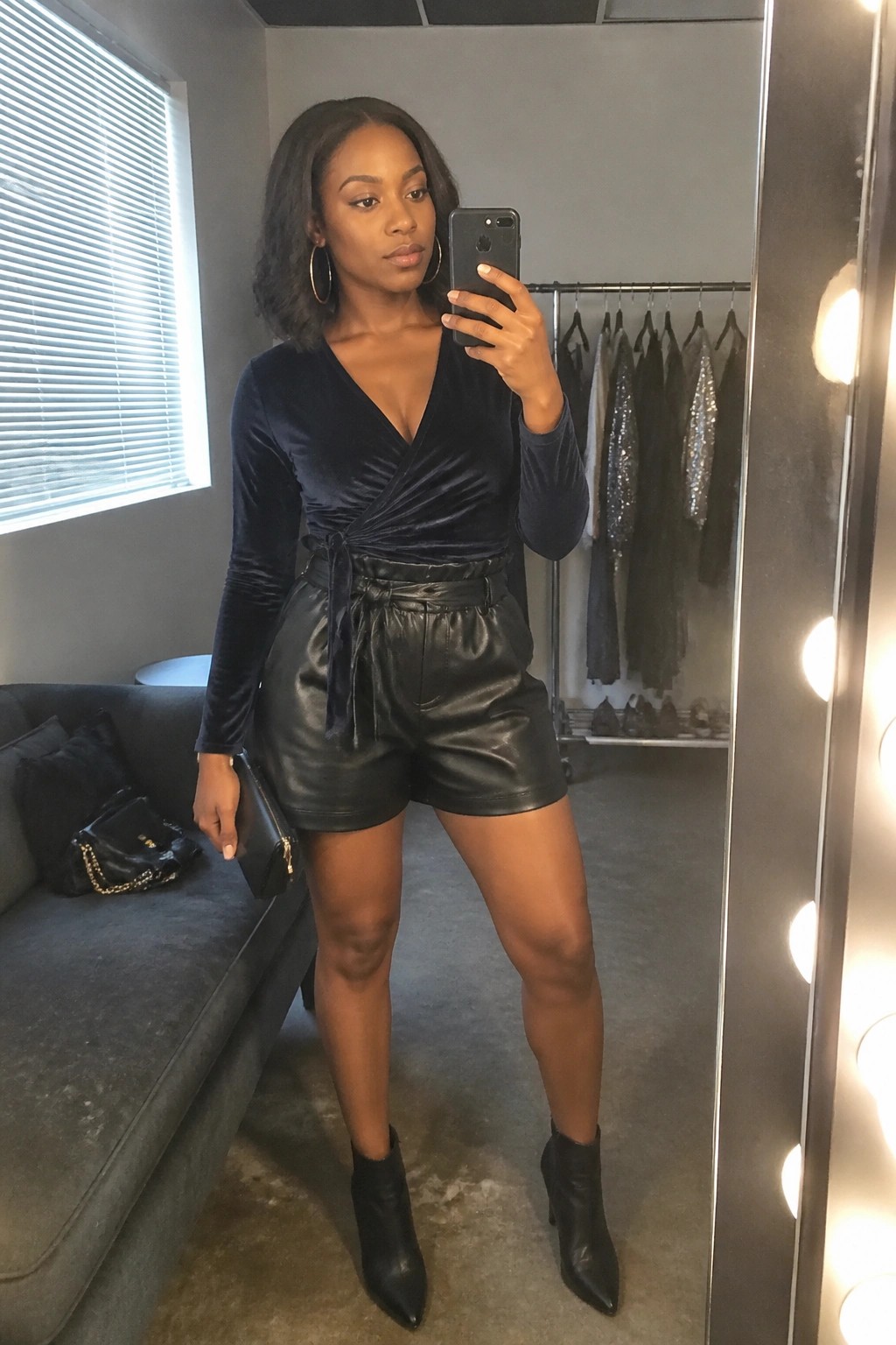 Woman in navy velvet wrap top and black leather shorts taking mirror selfie in boutique.
