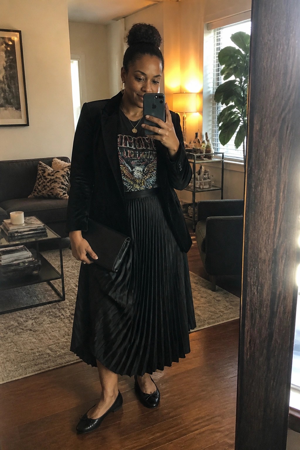 Woman in black blazer and pleated skirt taking mirror selfie in living room