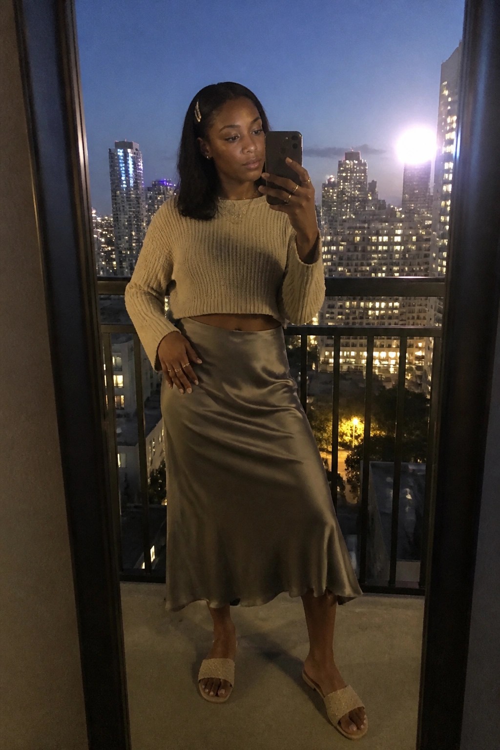 Woman wearing beige cropped knit sweater tucked into taupe satin midi skirt with beige woven slides and gold jewelry holding phone for mirror selfie.
