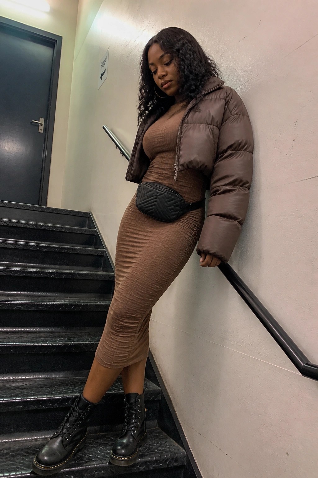 Woman in a cropped brown puffer jacket over a matching ruched midi dress paired with black combat boots and a black belt bag standing on stairs.