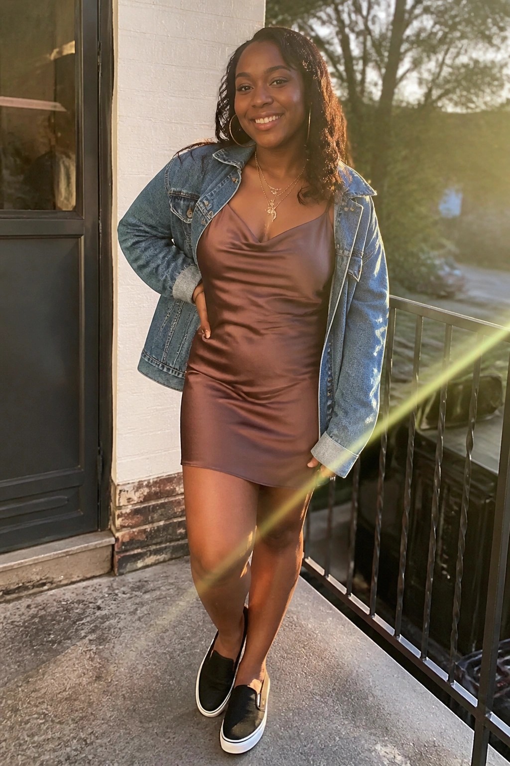 Woman in brown satin dress and denim jacket smiling on sunny balcony