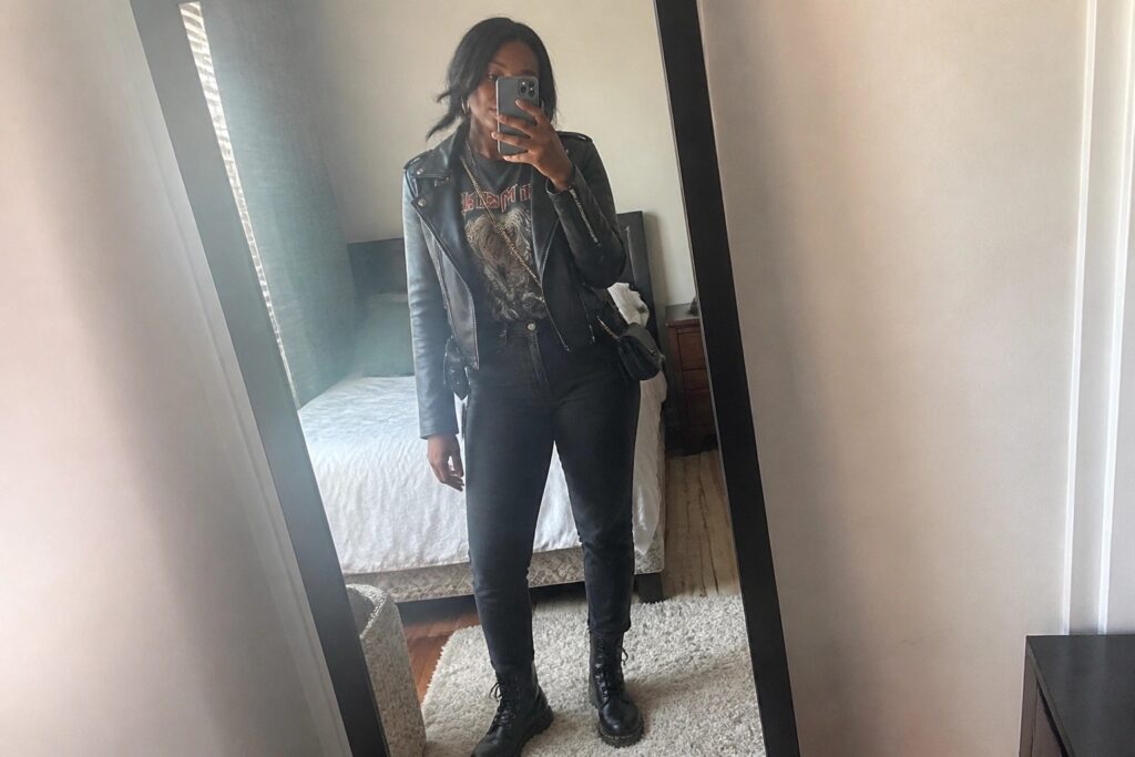 19 Elevated Concert Outfit Ideas for Black Women Without Heels Woman wearing black cropped leather jacket over graphic tee with chain necklace black jeans and lace up combat boots in mirror selfie.