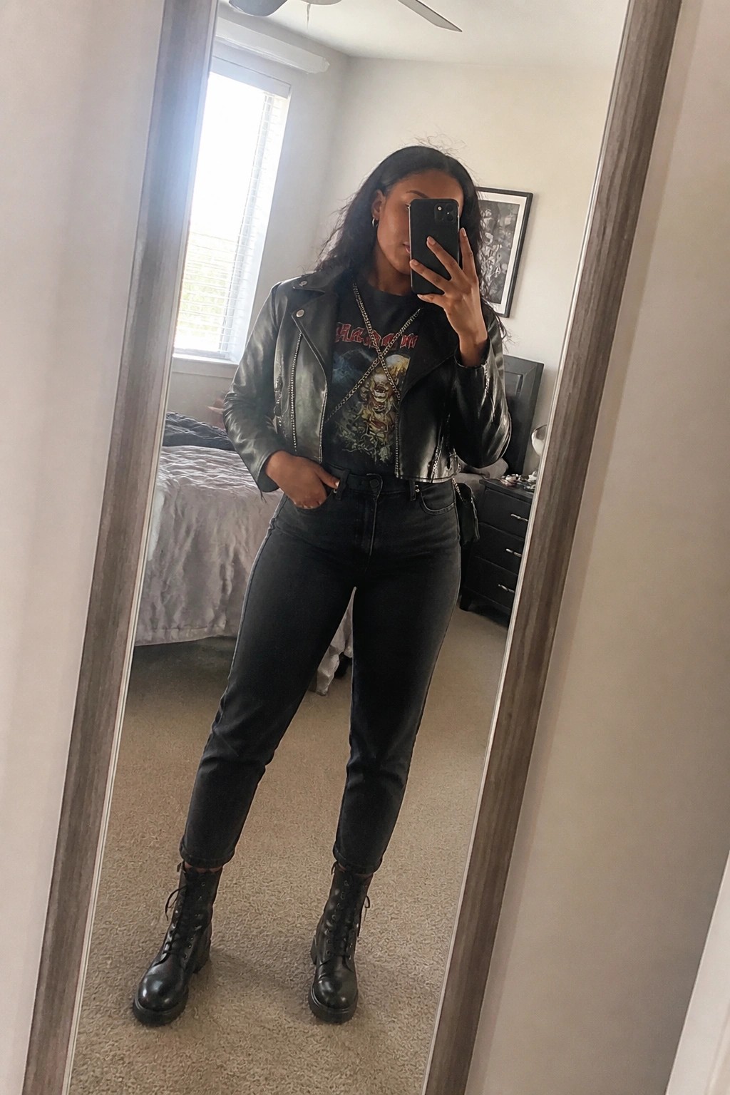 Woman wearing black cropped leather jacket over graphic tee with chain necklace black jeans and lace up combat boots in mirror selfie.