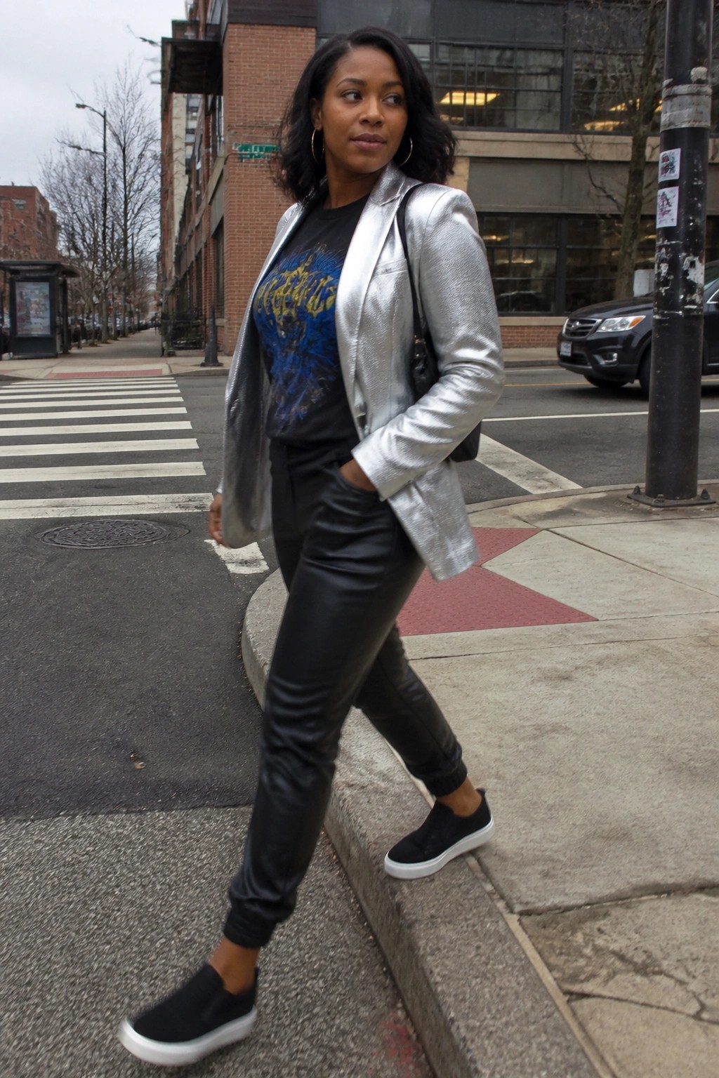 A Black woman in a silver metallic blazer layered over a black graphic t-shirt with blue and yellow print, black faux leather pants, and black slip-on sneakers with white soles, carrying a black shoulder bag on a city sidewalk.