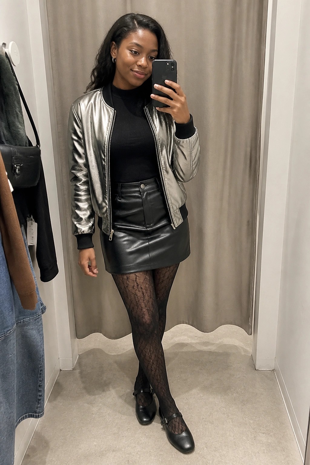 Woman wearing a silver metallic bomber jacket over a black turtleneck, paired with a black leather mini skirt, patterned black tights, and black mary jane flats in a fitting room mirror selfie.