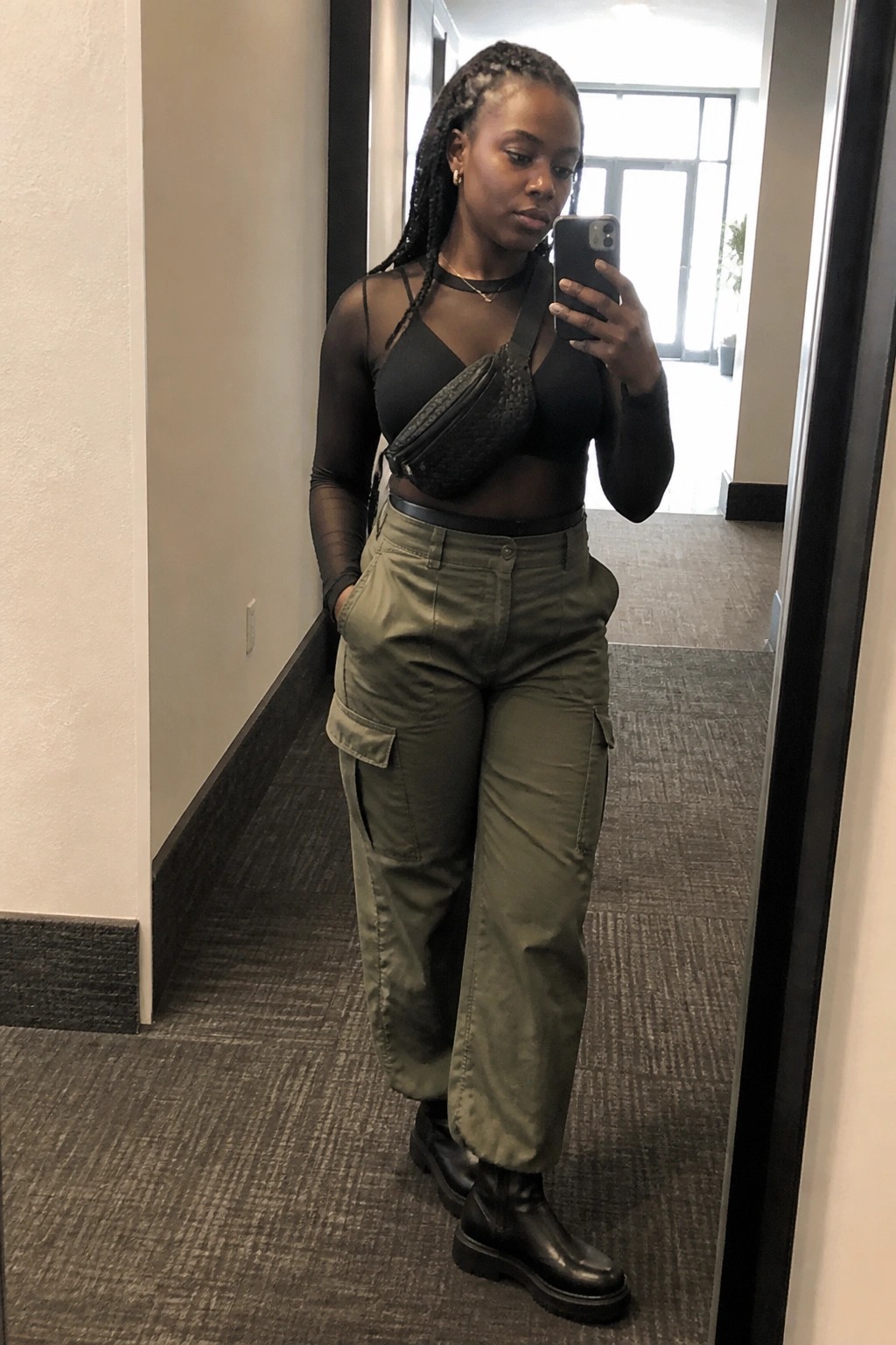 Woman in sheer black top and green cargo pants taking mirror selfie with phone.