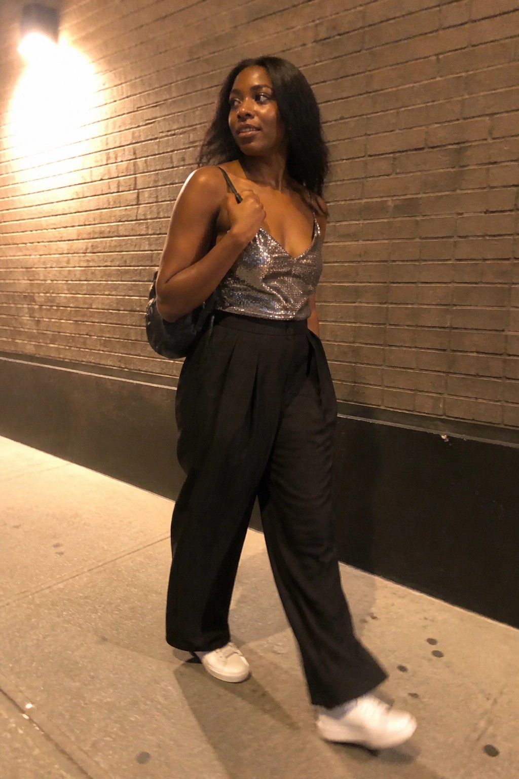 Woman in silver sequin top and black pants by brick wall at night